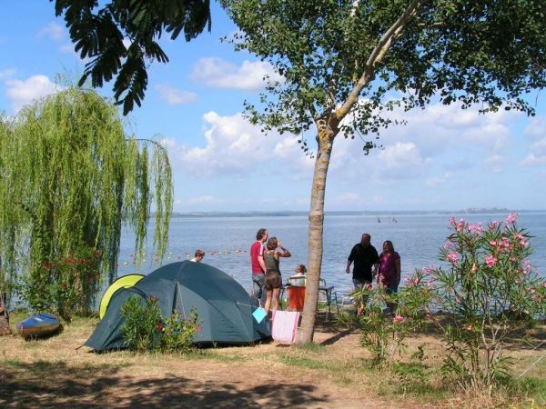 Una tenda da campeggio vicino a un lago, persone in piedi e un cielo soleggiato.