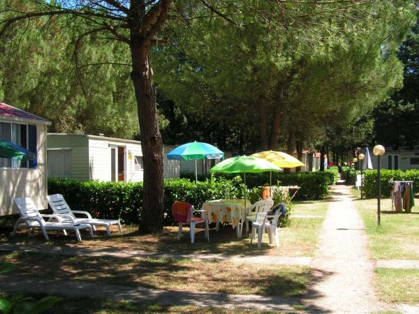 Percorso in un campeggio con case mobili, tavoli con sedie e ombrelloni colorati sotto gli alberi.