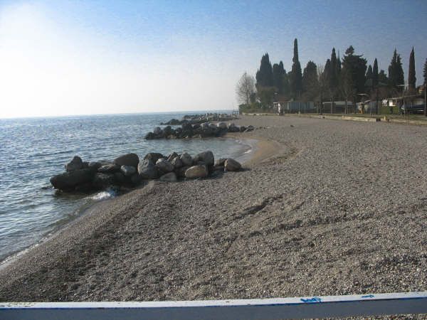Spiaggia di ciottoli con massi, lambita da un lago, circondata da alberi.