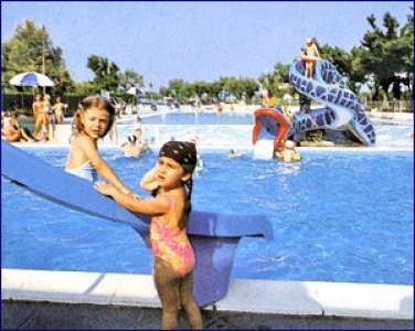 Bambine accanto a una piscina con scivolo, altre persone in acqua.