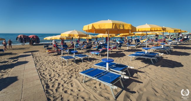 Spiaggia soleggiata con lettini blu e ombrelloni gialli allineati.